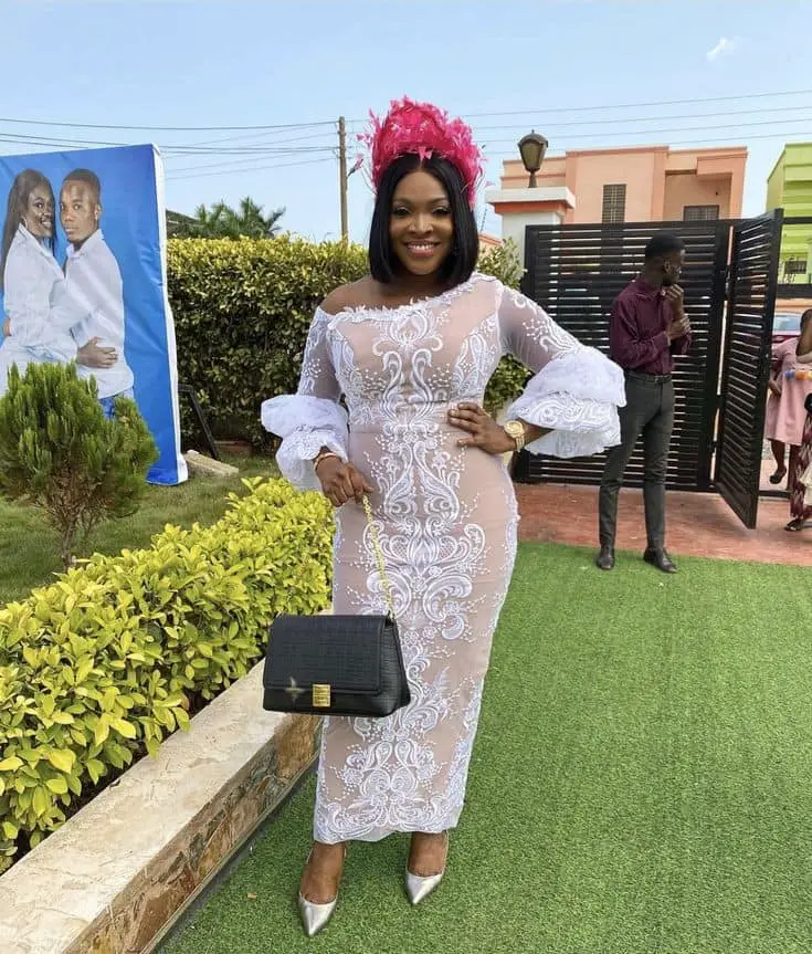 A lady in wedding guest white lace dress styles and black bag in Ghana 